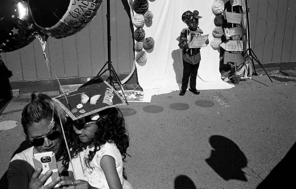 Black and White Photograph Depicting an elementary school graduation ceremony in East San Jose. A mother takes a selfie with her daughter in the lower left hand corder. Behind them a young graduate is getting her picture taken while holding up a graduation certificate.